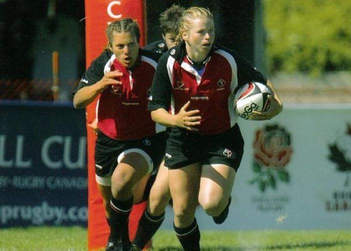 Two rugby players in red and black jerseys run across a field, one carrying a ball, near a red post and a sponsor banner.