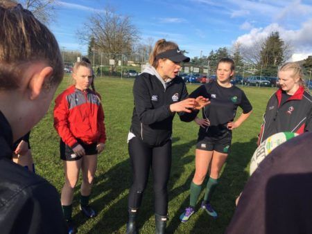 A coach in a black tracksuit gives instructions to a group of athletes on a grassy field during a sunny day.