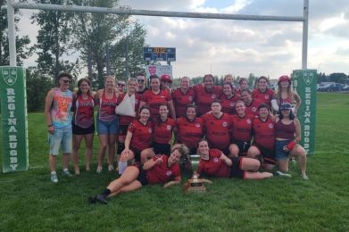 A rugby team in red jerseys posing with a trophy on a field between two goal posts under a blue, cloudy sky.