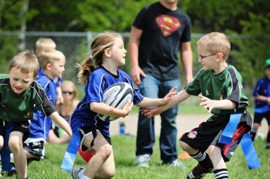 Children wearing team jerseys play a game of flag football on a grassy field, with one player carrying a rugby ball.