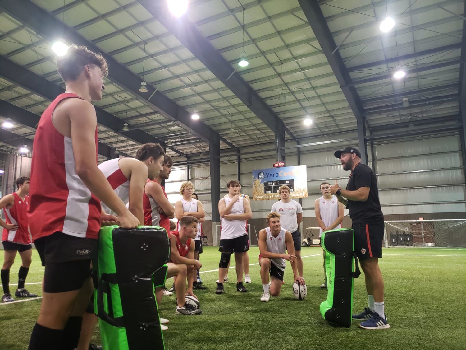 A coach speaks to a group of athletes gathered on an indoor turf field, standing near large green training pads.