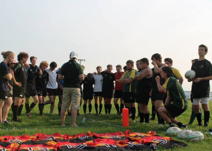 A coach addresses a group of rugby players standing in a circle on a field, with team jerseys laid out in the foreground.