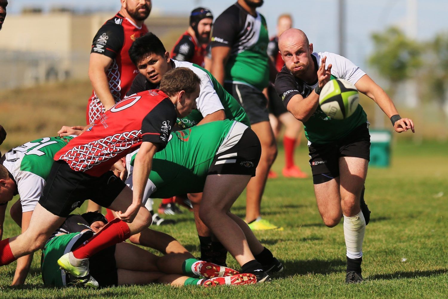 Rugby players in red and green jerseys compete on a grass field, with one player running forward while holding the ball.
