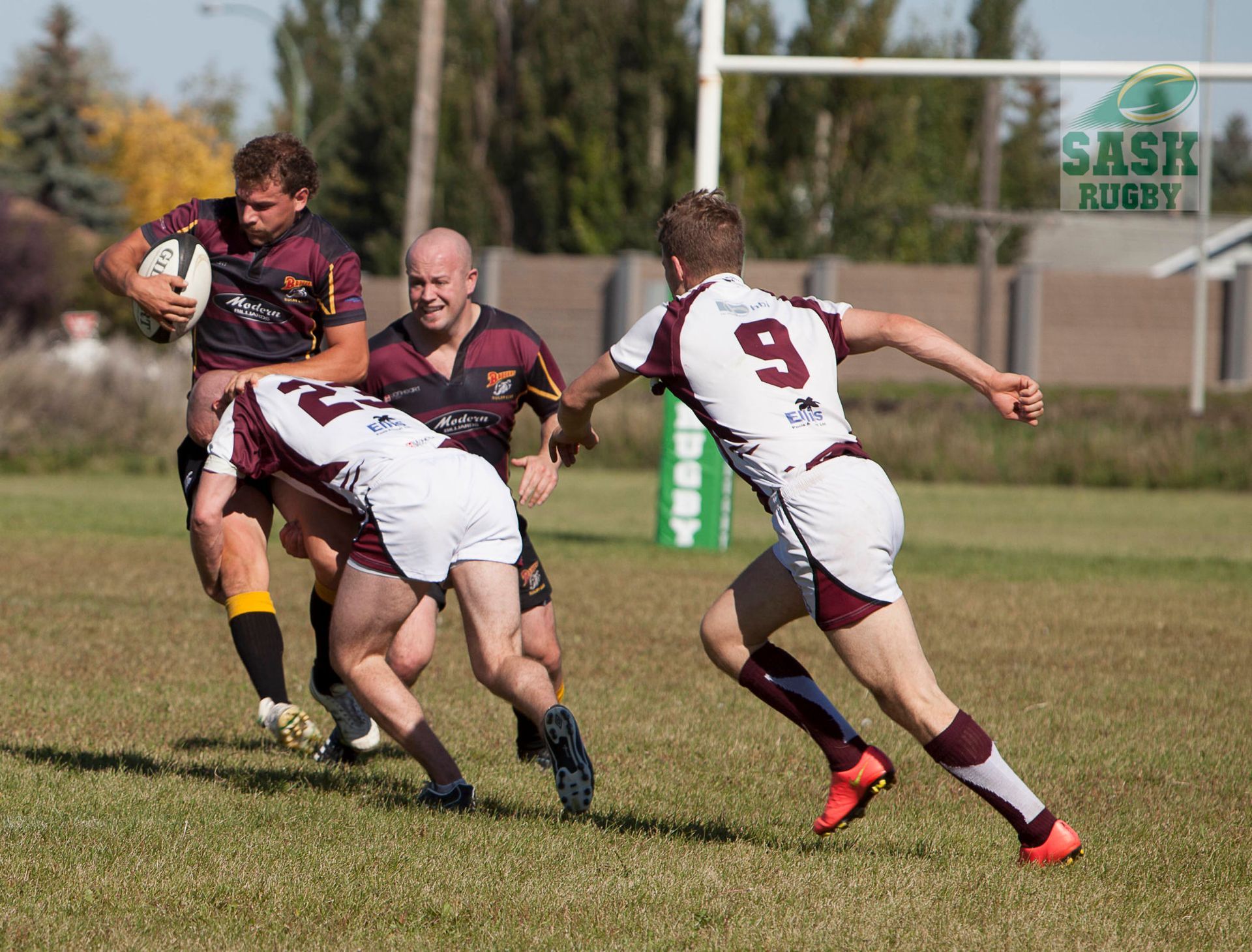 Rugby players in maroon and white uniforms compete for the ball on a sunny grass field.