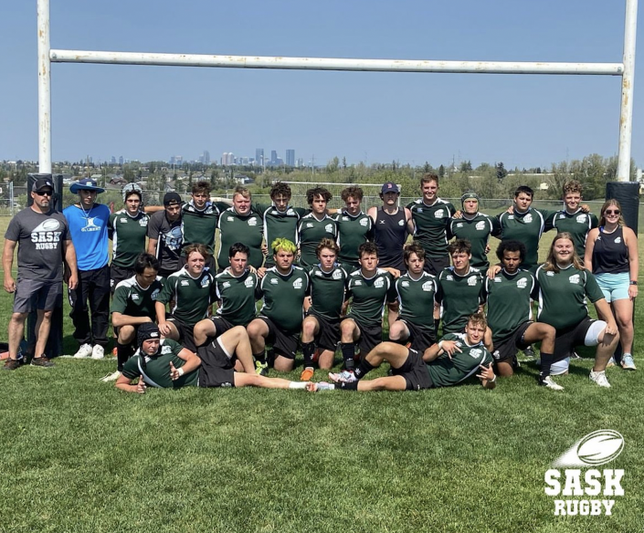 A rugby team wearing green and white jerseys poses for a group photo on a grassy field in front of rugby goalposts.
