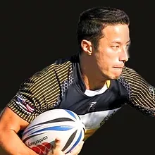 A rugby player in a black and yellow striped jersey holds a rugby ball while looking to the side against a dark background.