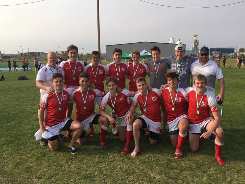 A team of players in red jerseys and white shorts with medals poses on a grass field, with two coaches standing behind.