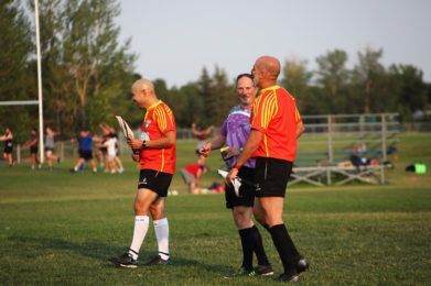 Three officials in bright jerseys and shorts walk and talk on a grassy sports field during a game.