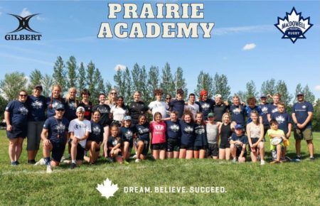 A large group of athletes and coaches pose on a grass field under a sunny sky for a Prairie Academy team photo.