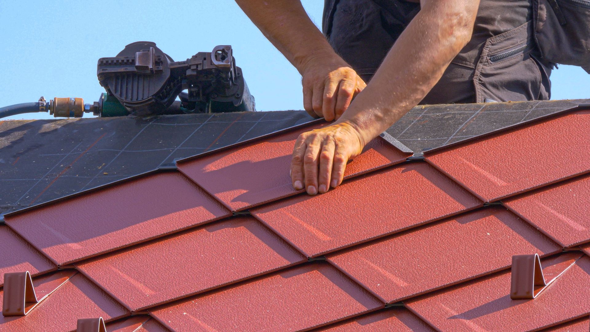 Worker installing red roof shingles on a house roof with a nail gun nearby.
