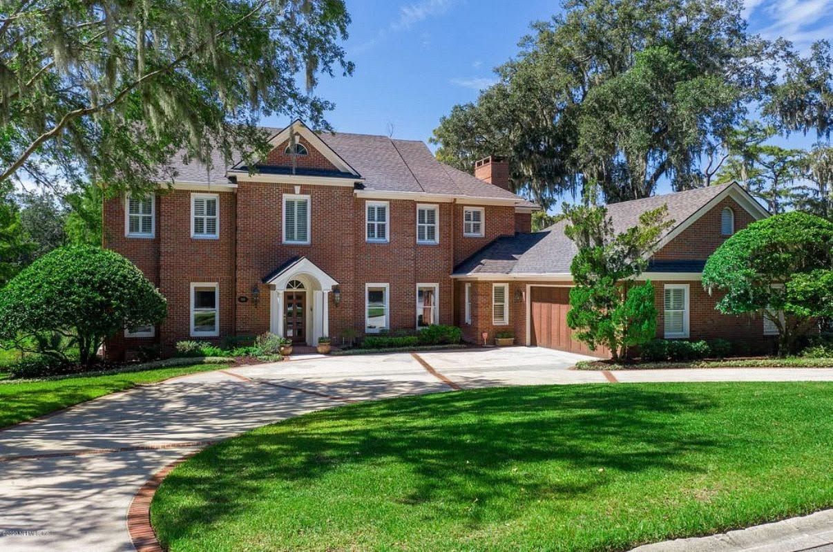 Large brick suburban house with a curved driveway, green lawn, and trees on a sunny day