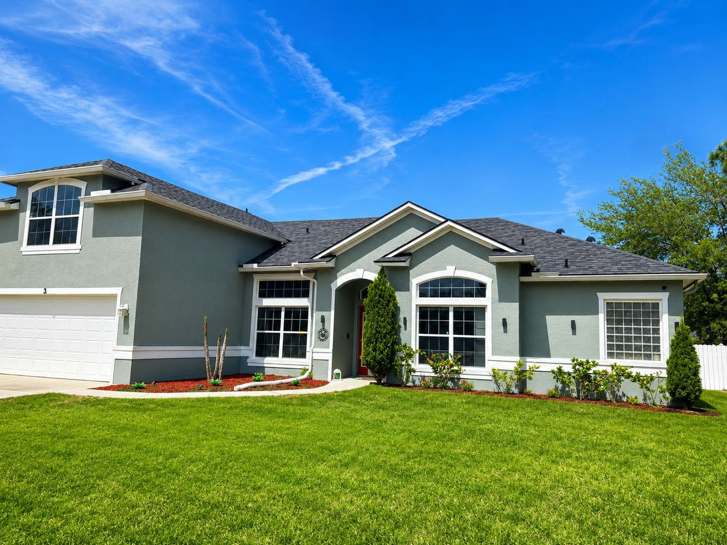 Single-story suburban house with gray exterior, white trim, and bright green lawn under a blue sky