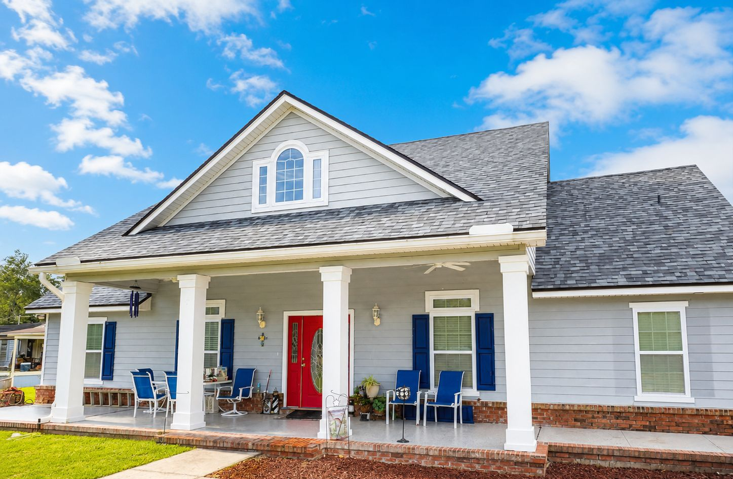 White house with blue shutters, red front door, and covered porch with blue chairs under a bright sky