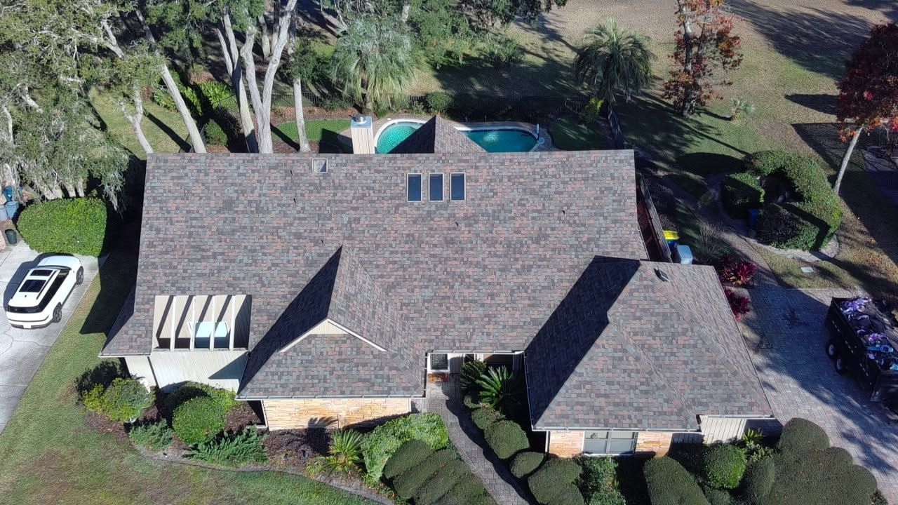 Aerial view of a house with a dark roof, skylights, and surrounding trees and driveway.