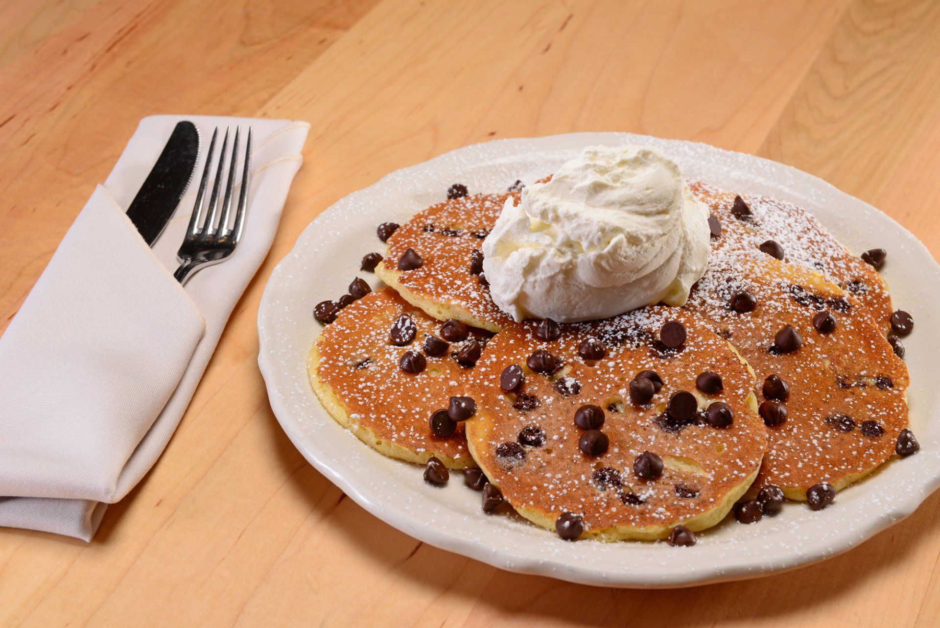 Chocolate chip pancakes with whipped cream, on a plate with silverware and napkin.