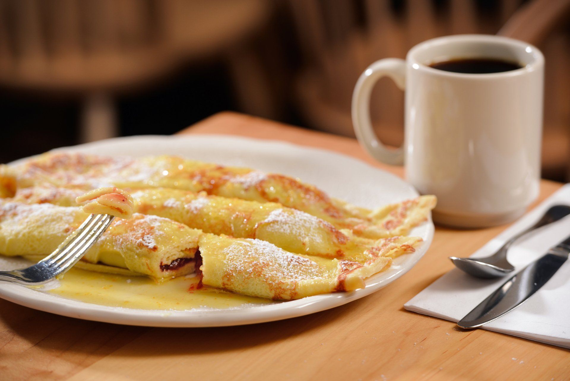 Crepes dusted with powdered sugar on a plate with coffee, fork, and cutlery.