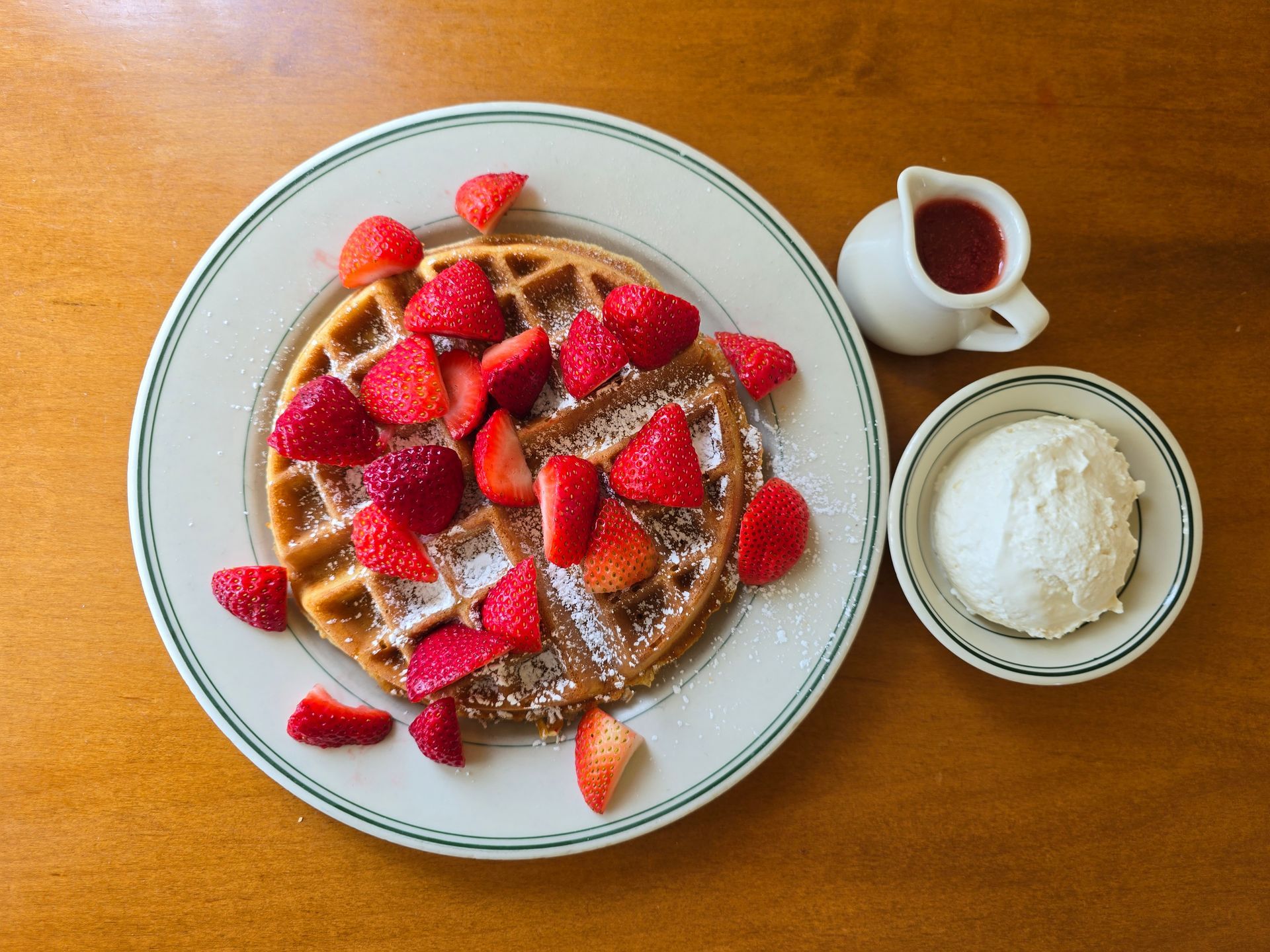 Waffles topped with strawberries and powdered sugar, served with cream and strawberry syrup.