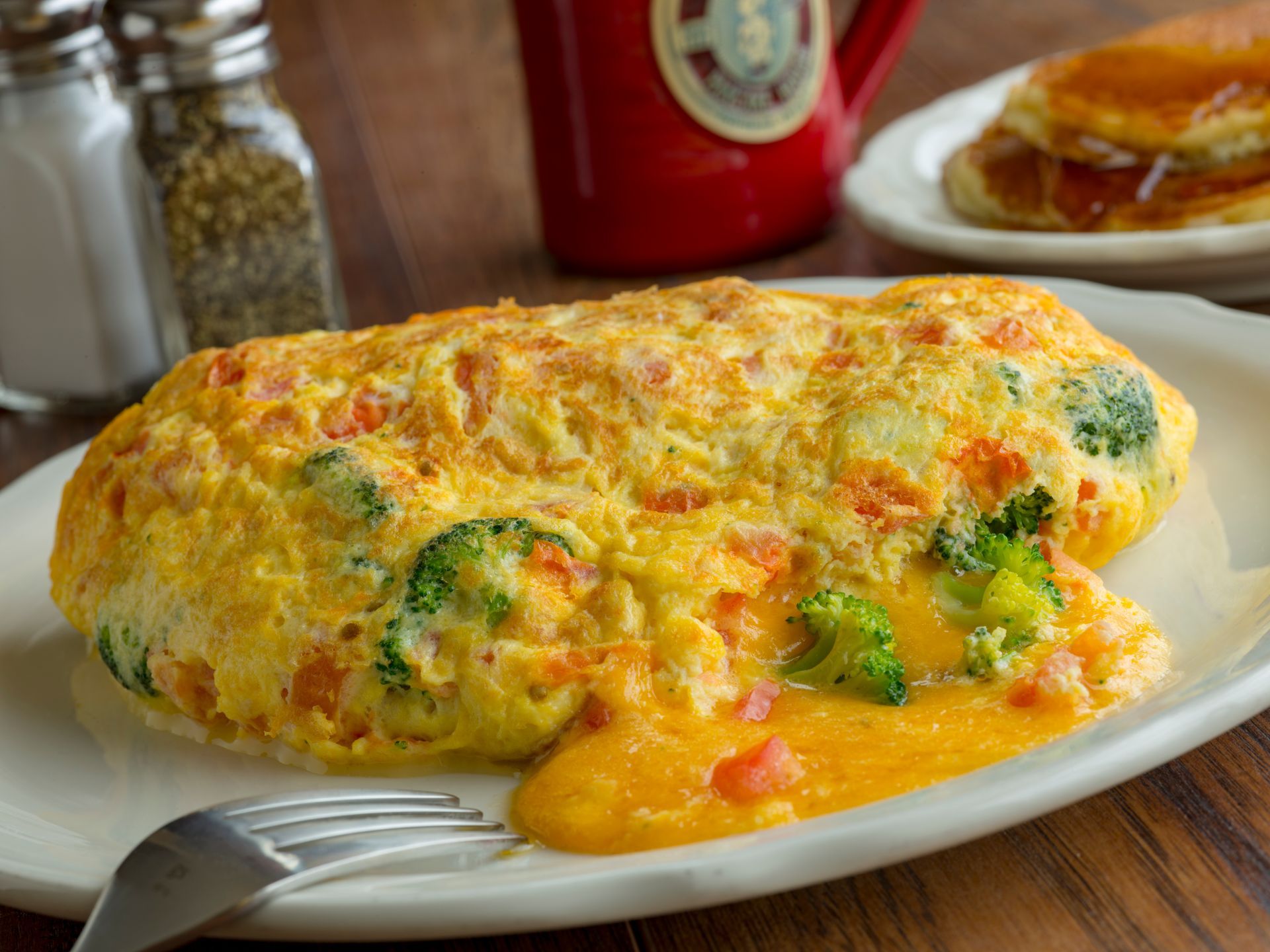 Omelet with broccoli, carrots, and cheese on a white plate. Pancakes and coffee in the background.