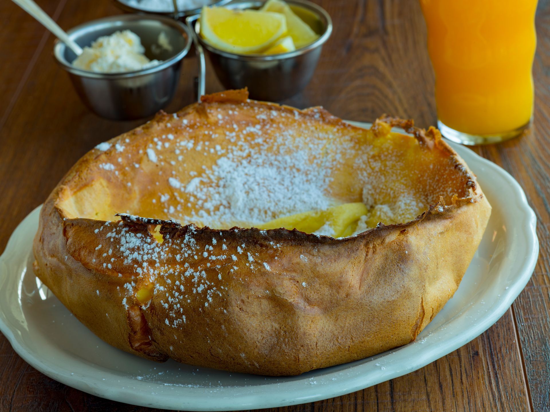 Dutch baby pancake dusted with powdered sugar on a white plate, served with lemon slices and orange juice.