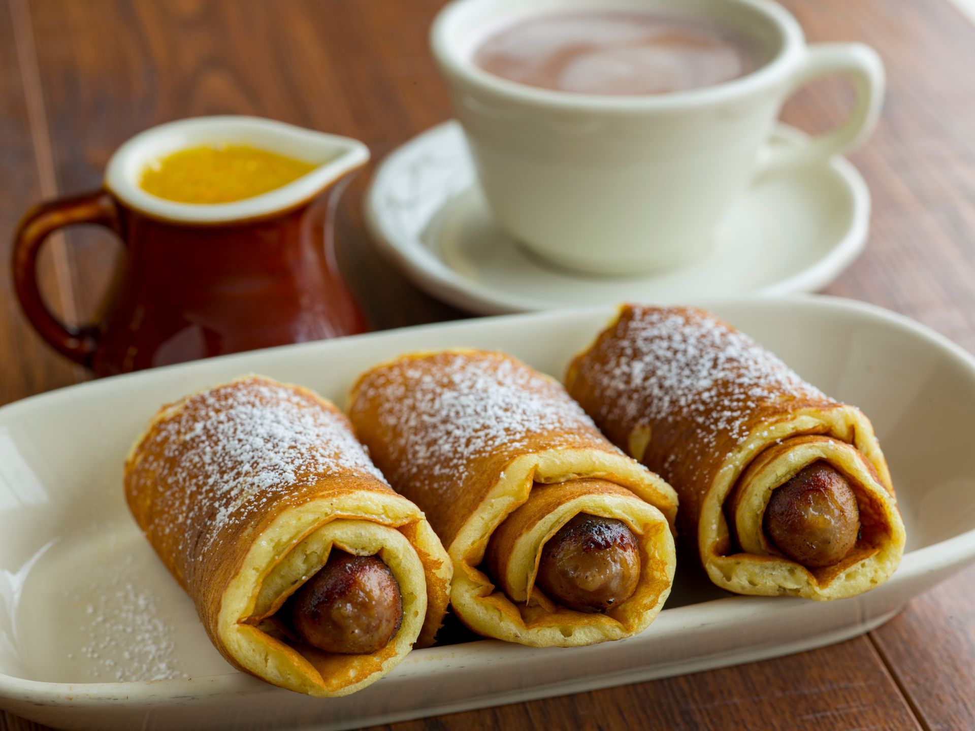 Sausage rolls dusted with powdered sugar on a platter, served with syrup and a mug of hot chocolate.