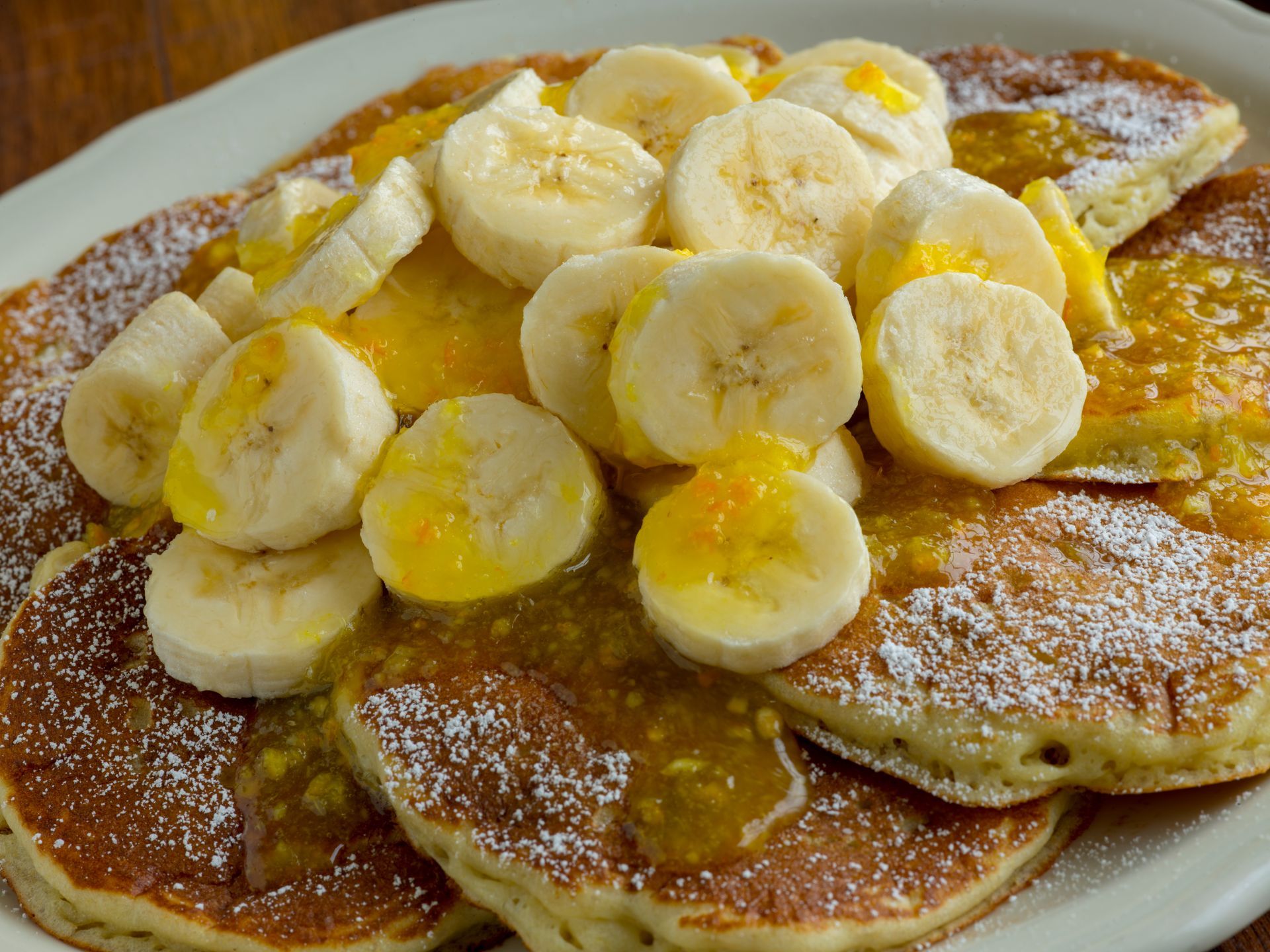 Pancakes with sliced bananas, orange sauce, and powdered sugar on a white plate.