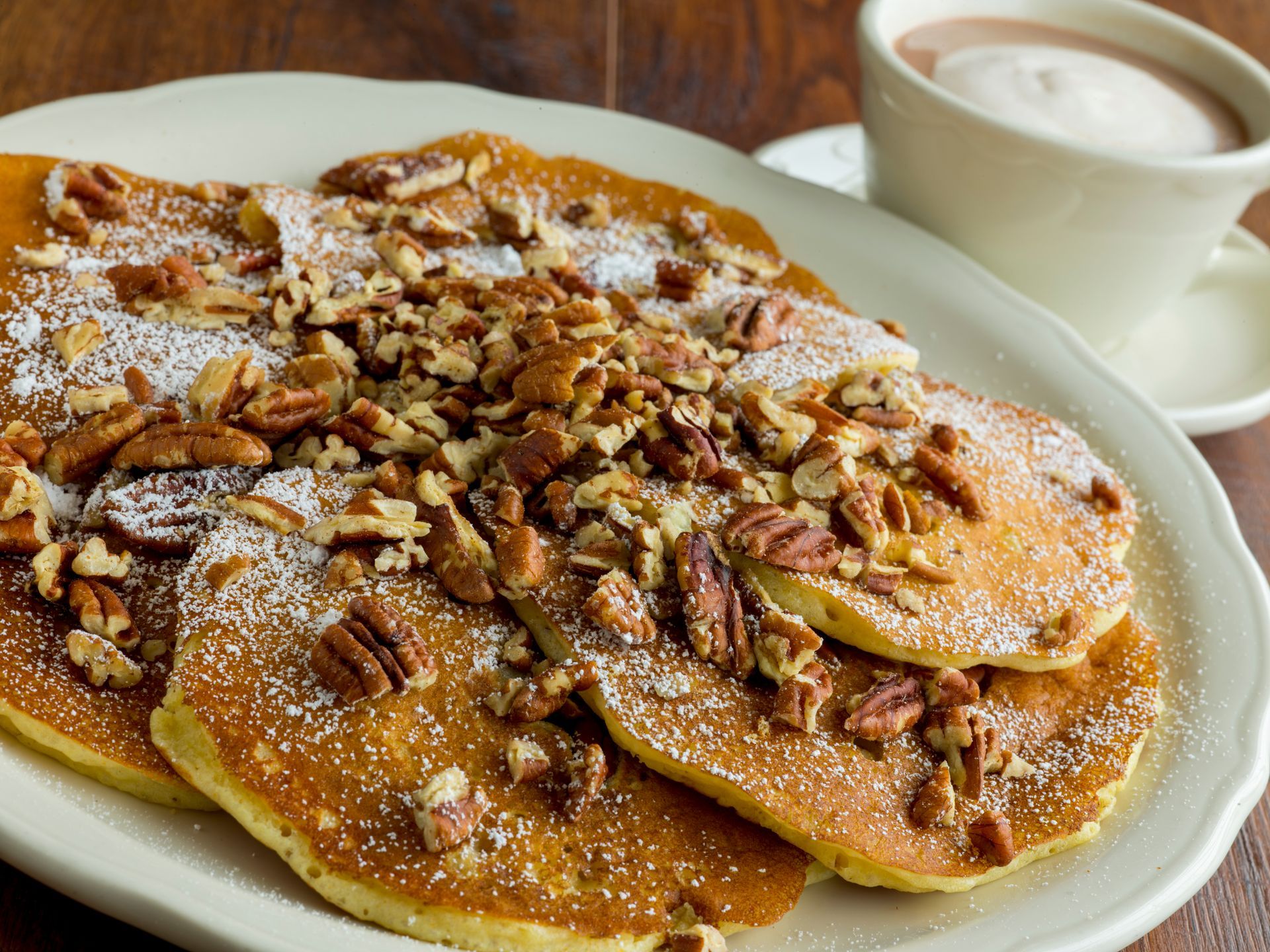 Pancakes topped with pecans and powdered sugar on a plate, with a cup of hot chocolate.