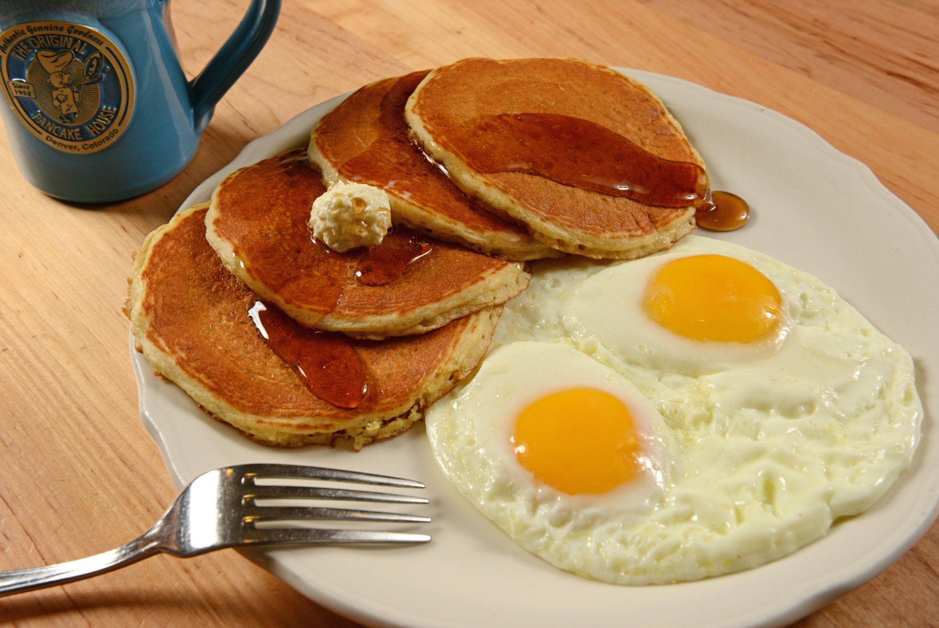 Breakfast plate with pancakes, syrup, butter, and two fried eggs; blue mug and fork.