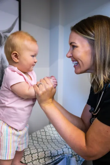 Adult smiling at a baby, holding hands in a bright room.