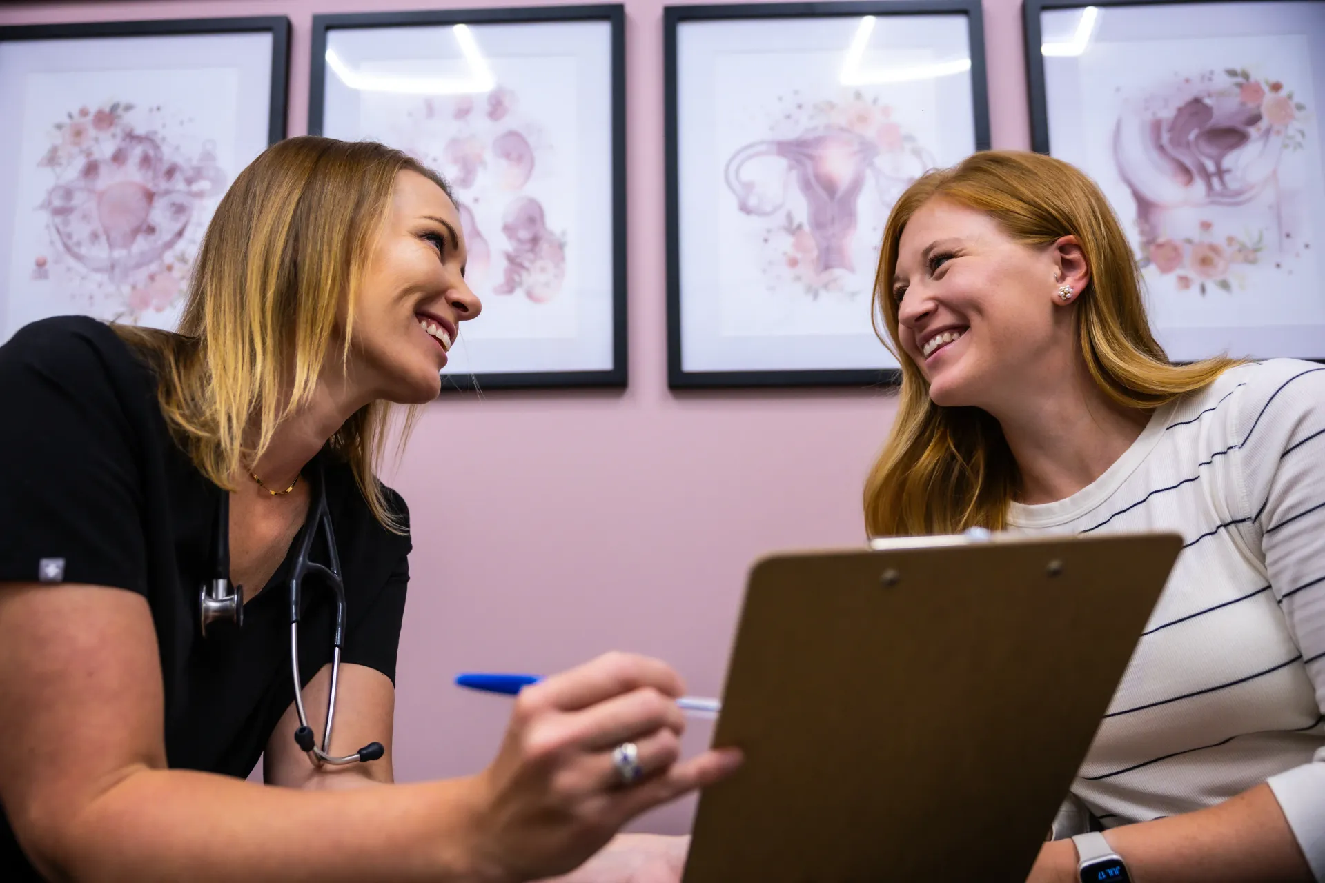 Two women smiling and talking across a table with a clipboard in a pink office.