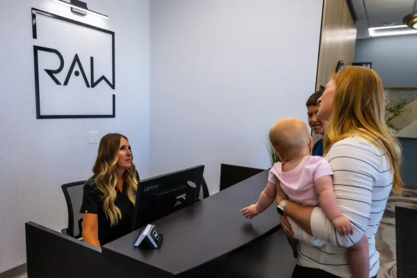 Reception desk with seated staff greeting two visitors, one holding a child, beside a RAW sign on the wall
