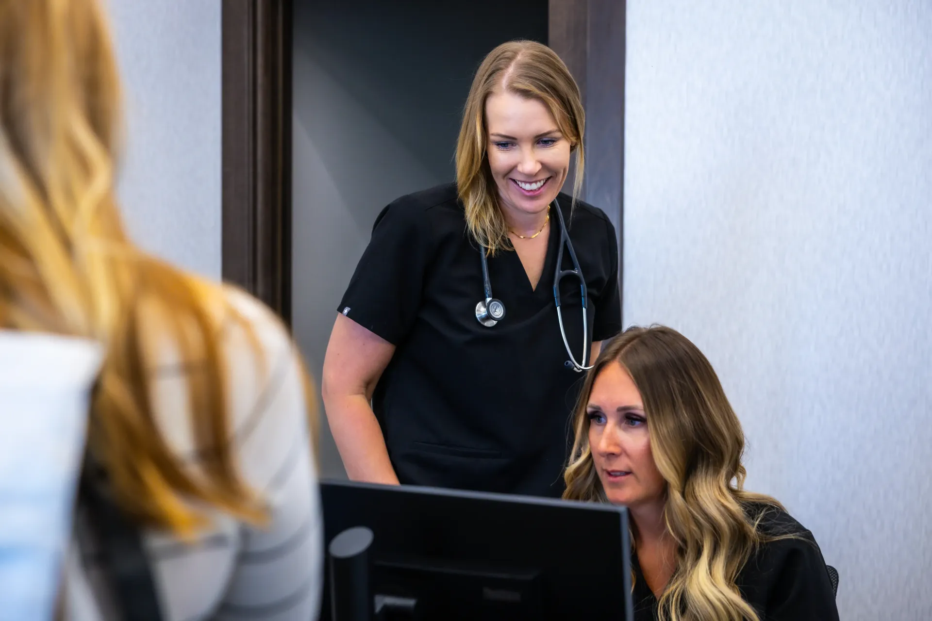 Two women in scrubs at a clinic desk, one smiling behind a computer while a patient stands nearby
