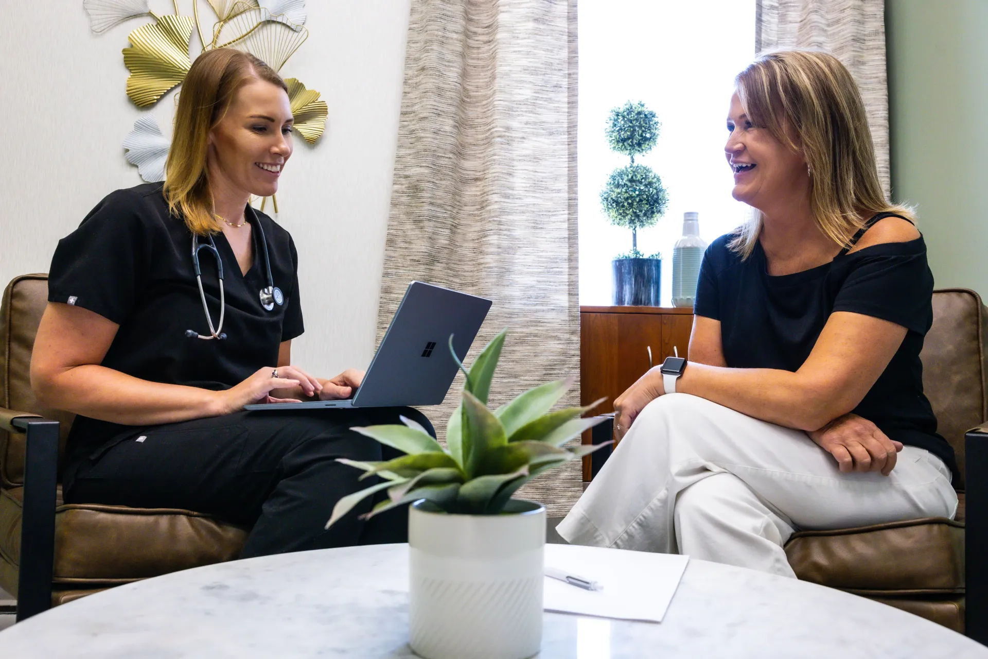 Two women chatting on couches in a bright office lounge, one using a laptop.