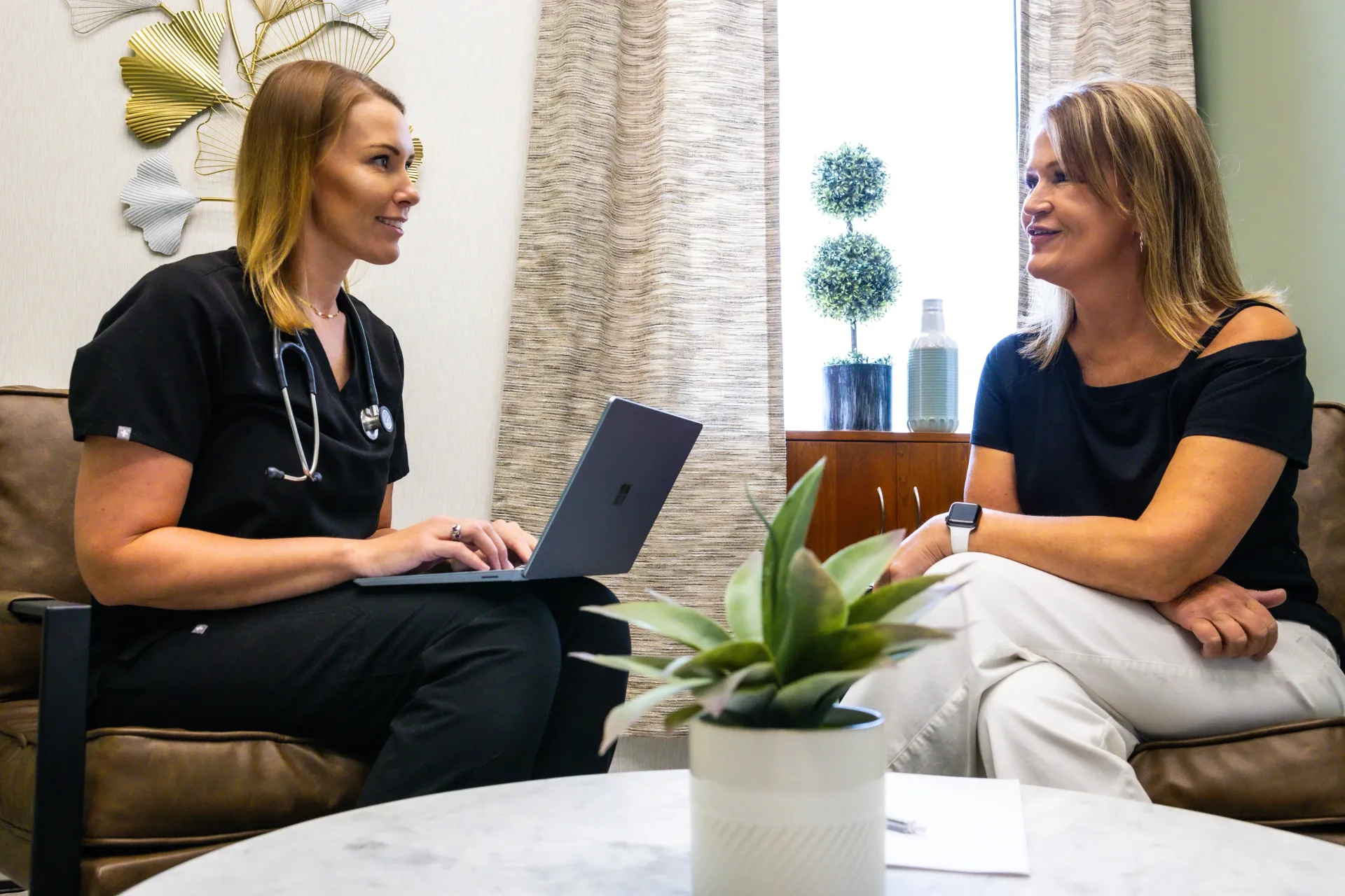 Two women chatting on a couch in a bright office lounge, one holding a laptop.