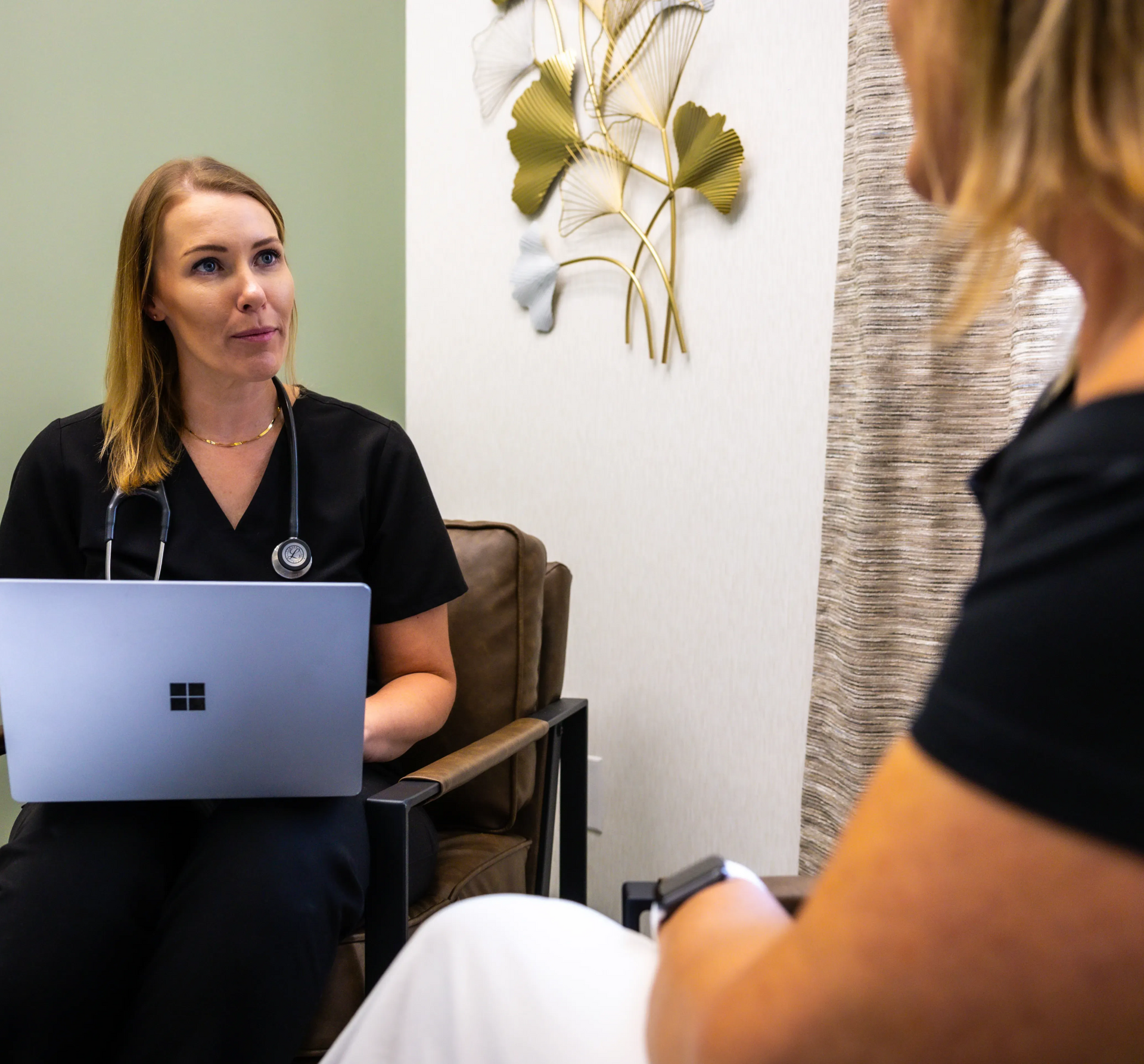 Therapist in black scrubs talks with a seated client in a calm office, laptop open on her lap.