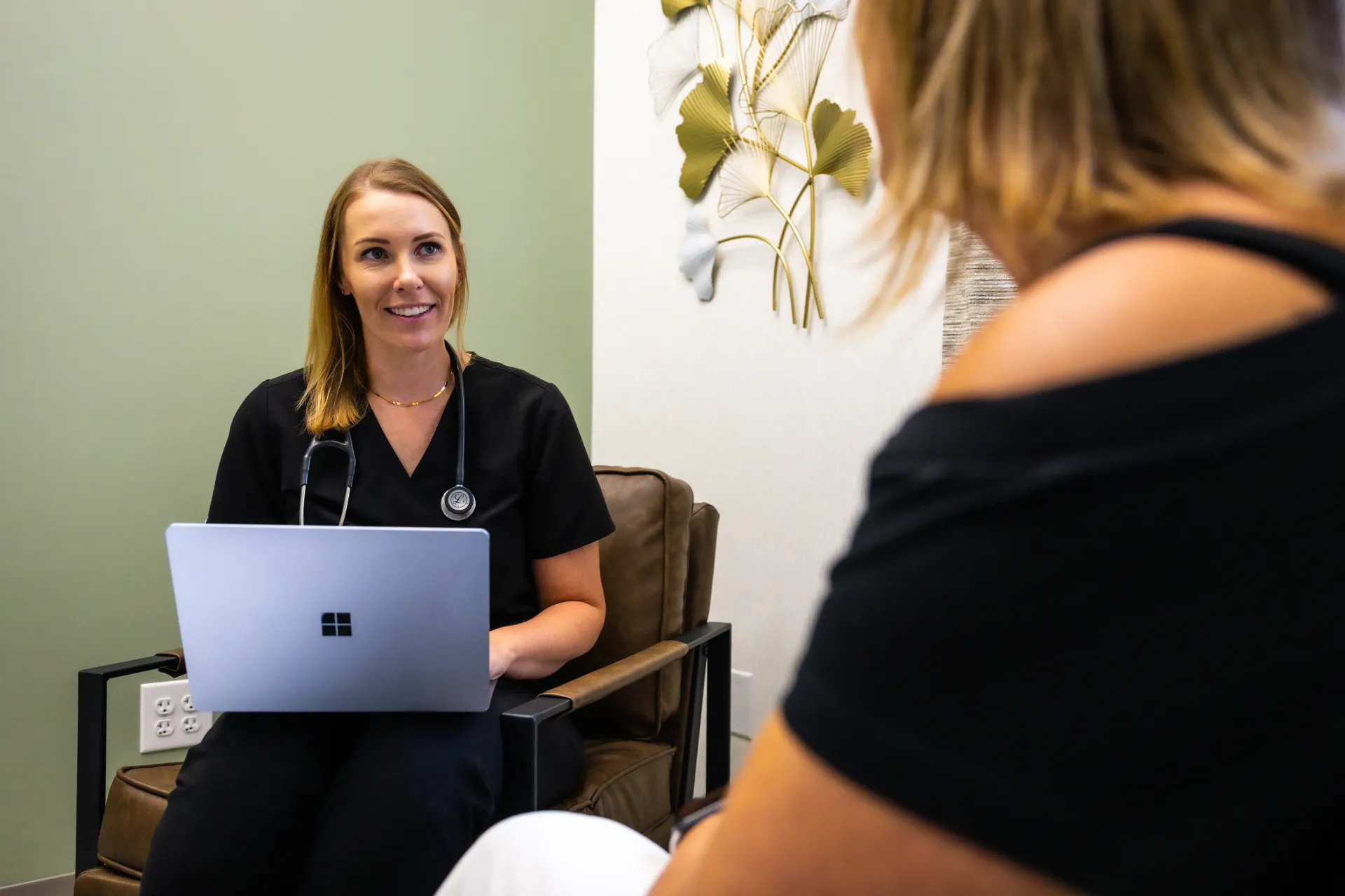 Two women in a consultation room; one speaks with a laptop while facing another seated client.