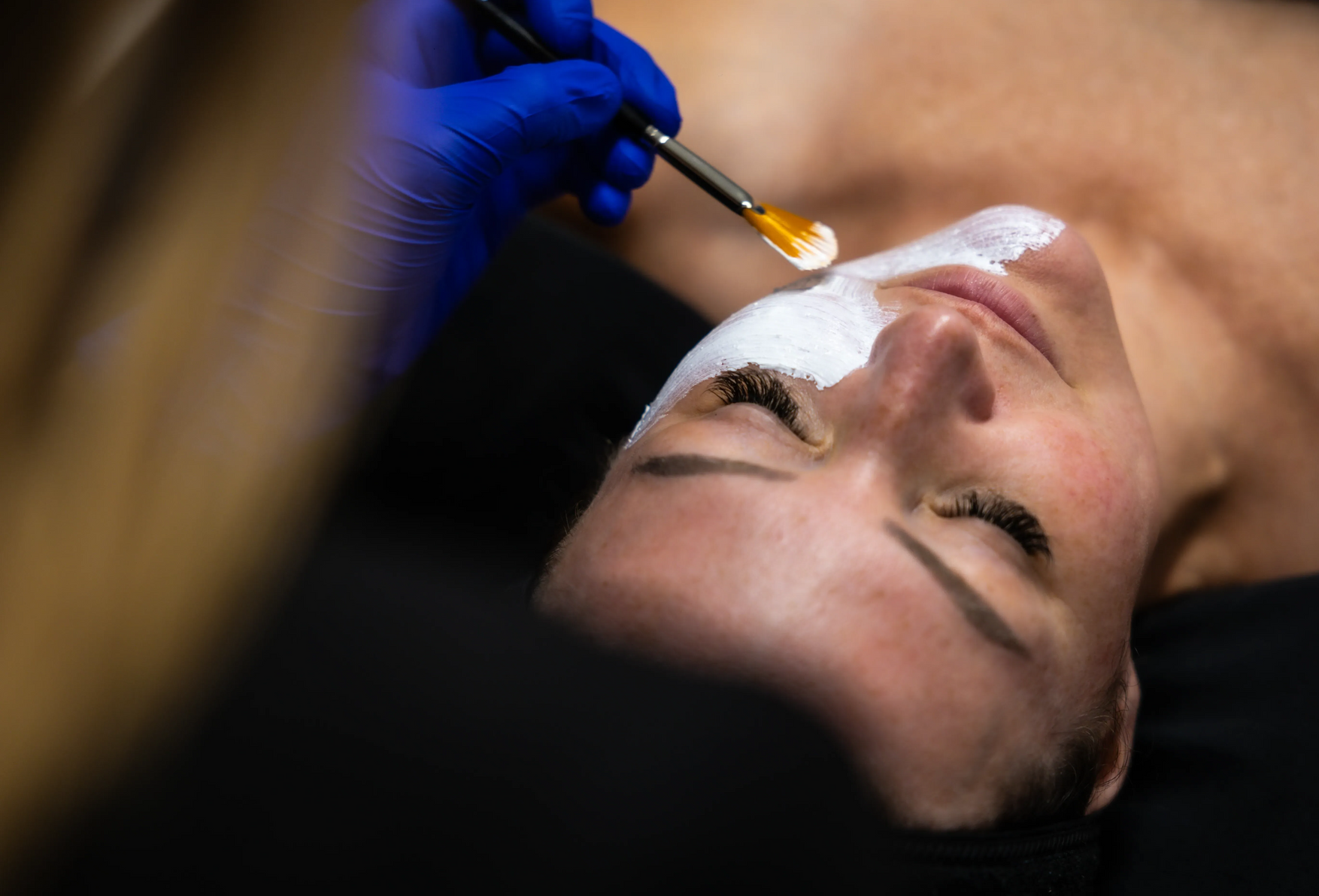 Esthetician applying gold facial serum with brush to a client lying on a treatment bed