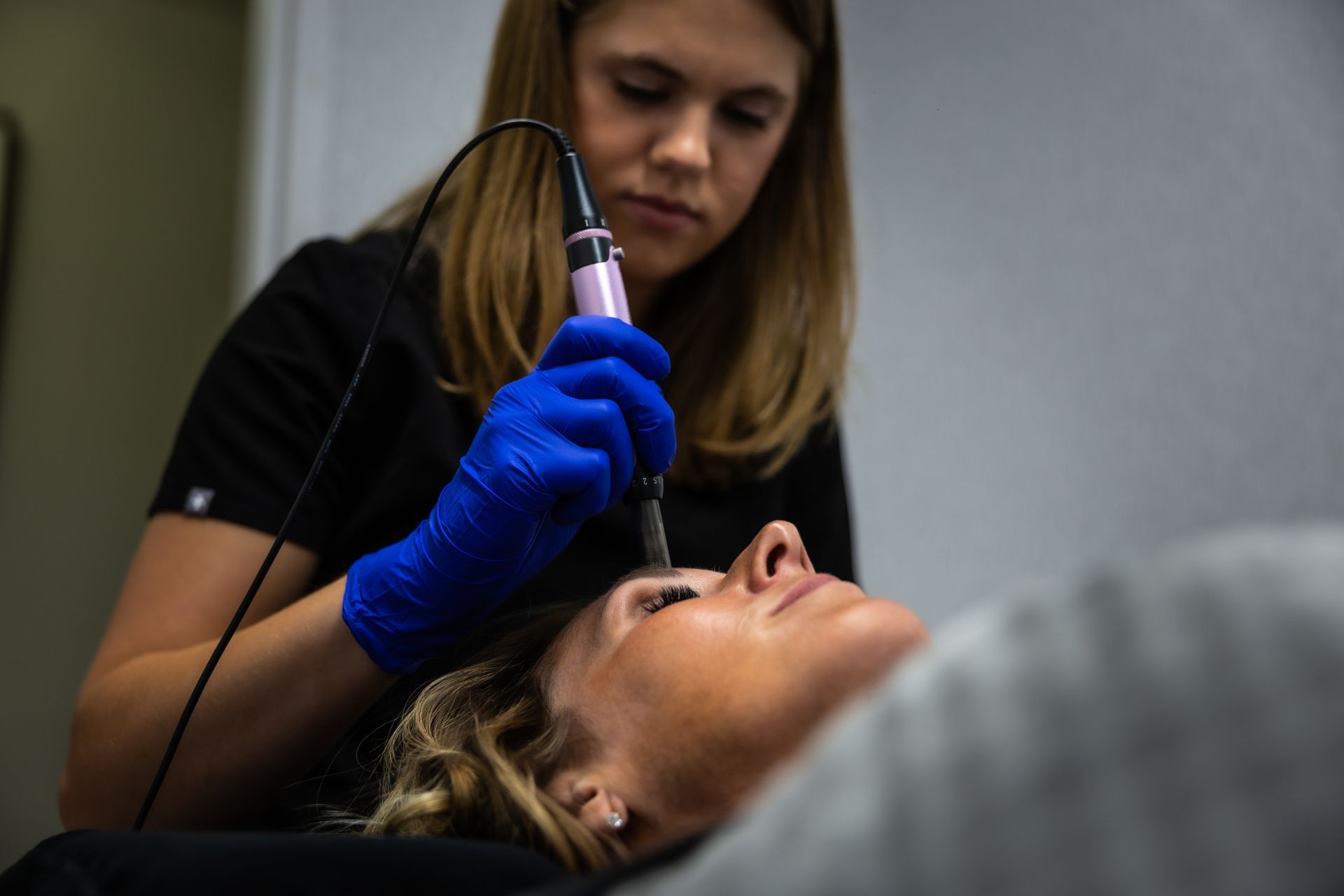 A clinician in blue gloves examines a patient’s face with a handheld device.