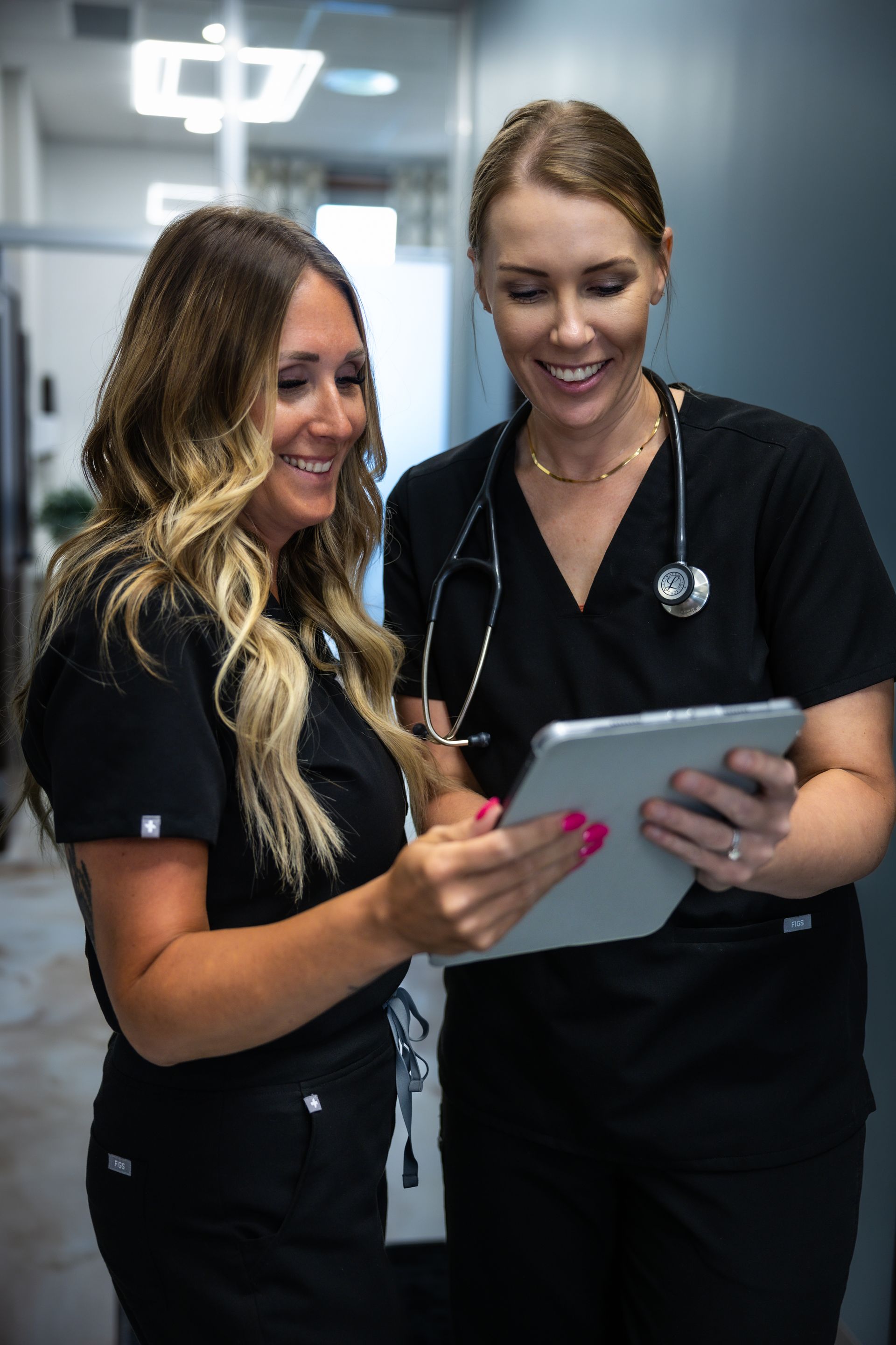 Two nurses in black scrubs smiling while looking at a tablet in a hospital hallway