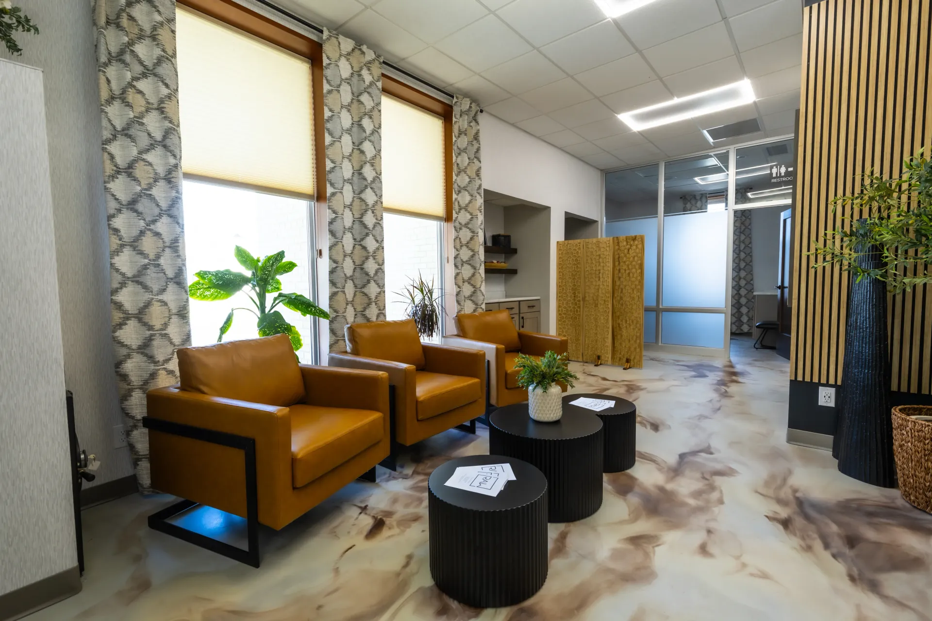 Modern waiting lounge with tan chairs, black tables, plants, and patterned walls in a bright office lobby