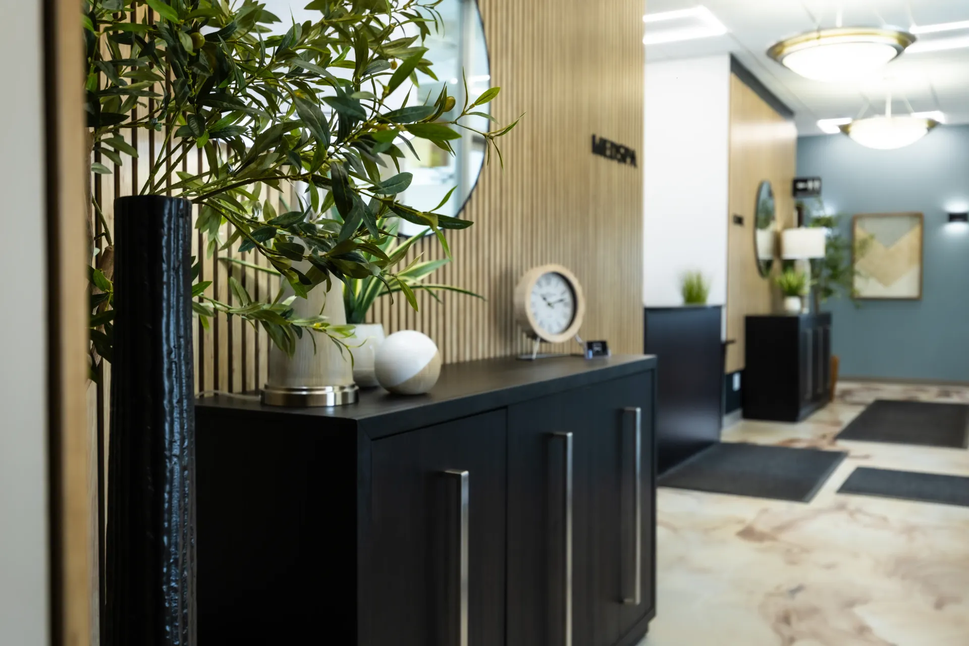 Modern office lobby with black cabinets, tall potted plants, wood paneling, and a hallway leading back