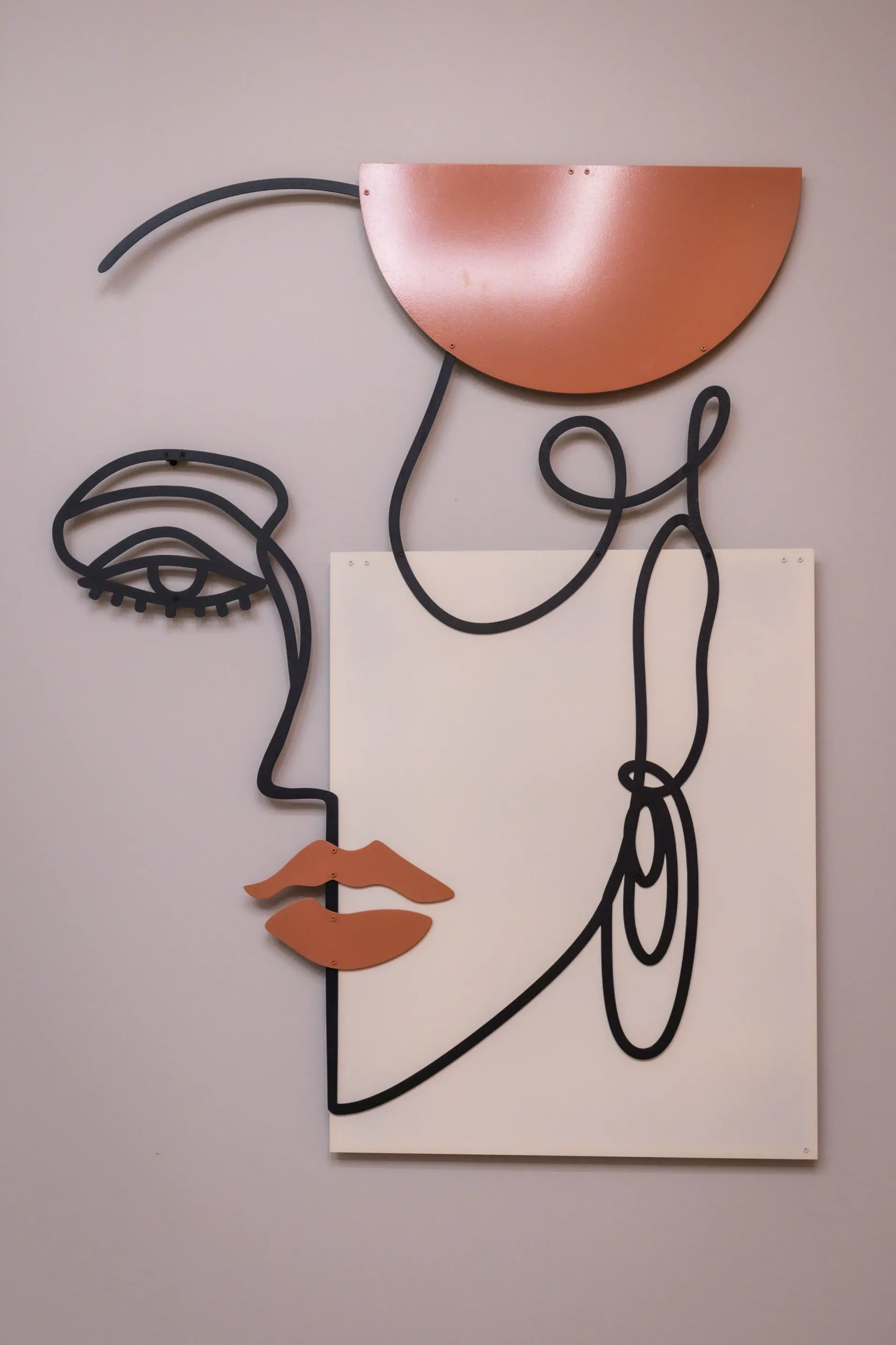 Abstract wall art of a face in black linework with copper semicircle and orange lips on a white background