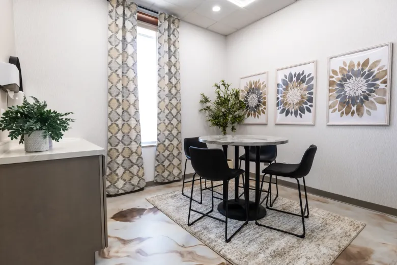 Small dining nook with a round black table, two chairs, floral wall art, and a potted plant.