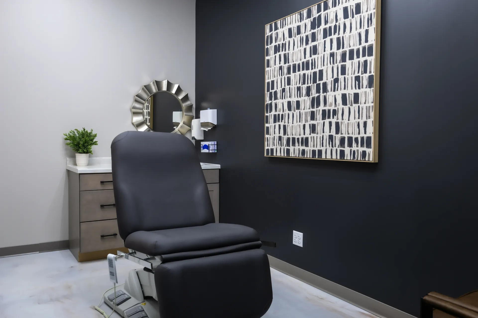 Modern exam room with black exam chair, dark wall, abstract art, and a small cabinet with plant.