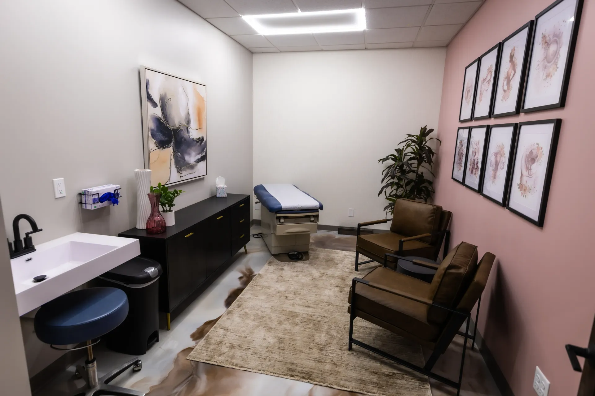 Small medical exam room with exam table, sink, chairs, and framed wall art on pink walls.