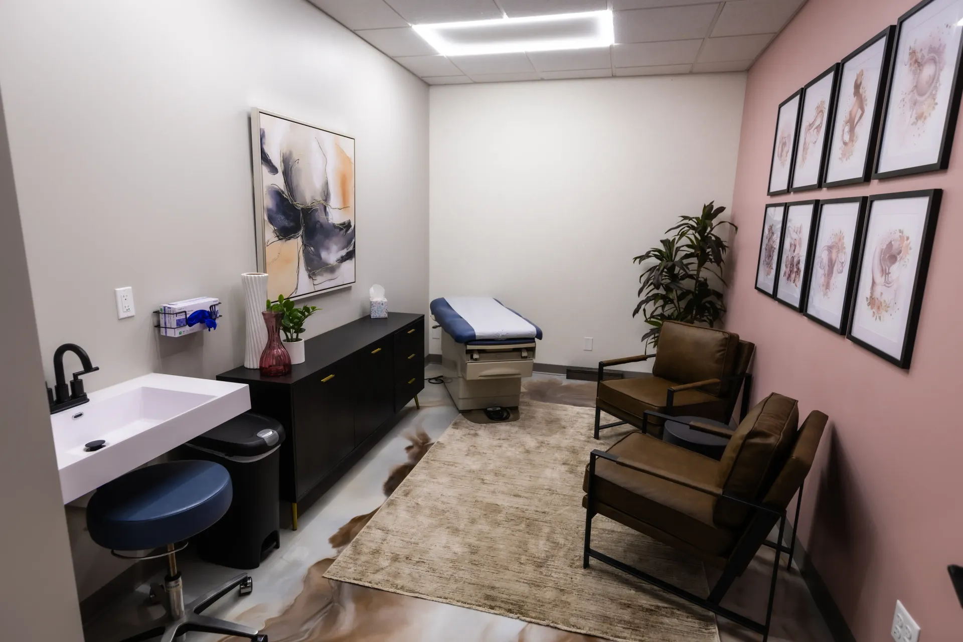 Small medical exam room with exam table, sink, chairs, and framed wall art.