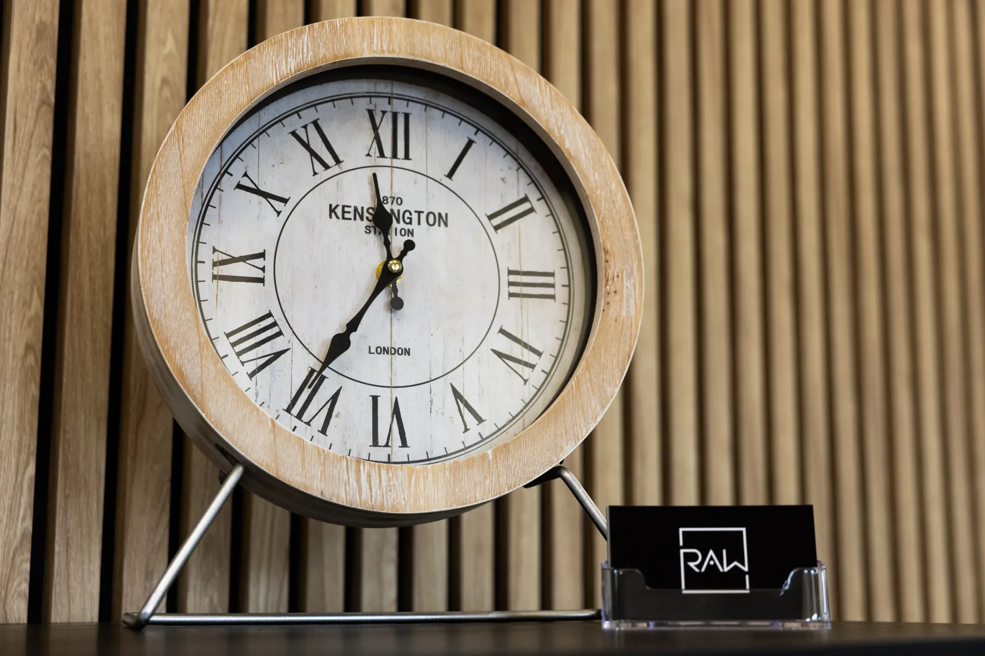 Decorative gold clock on a black stand beside a small nameplate against vertical wood slats.