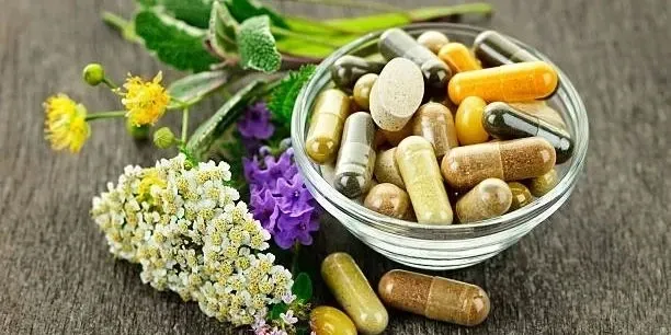 Assorted herbal capsules in a glass bowl with flowers and green leaves on wood surface