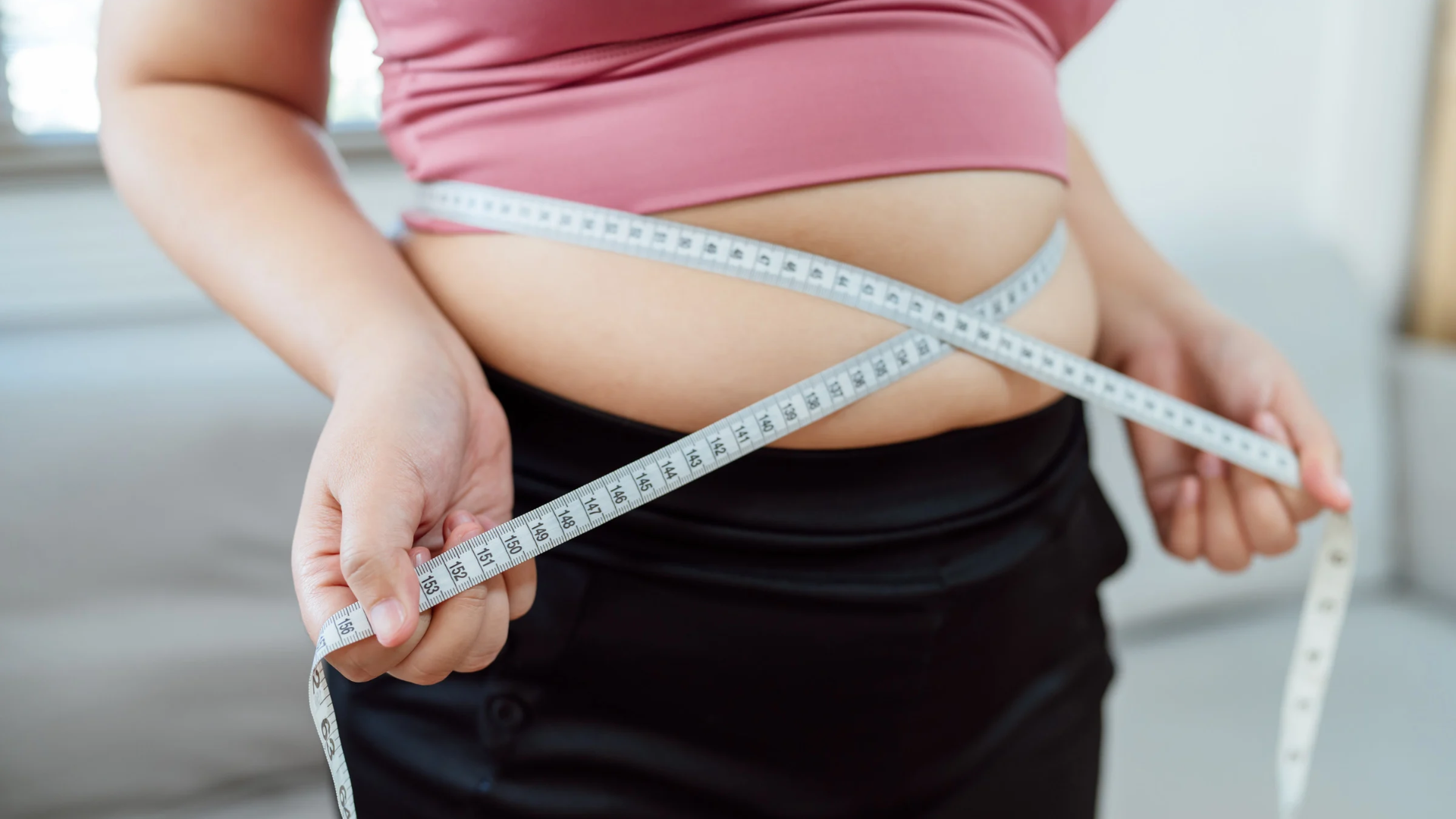 Person measuring their waist with a tape measure over a pink top and black pants.