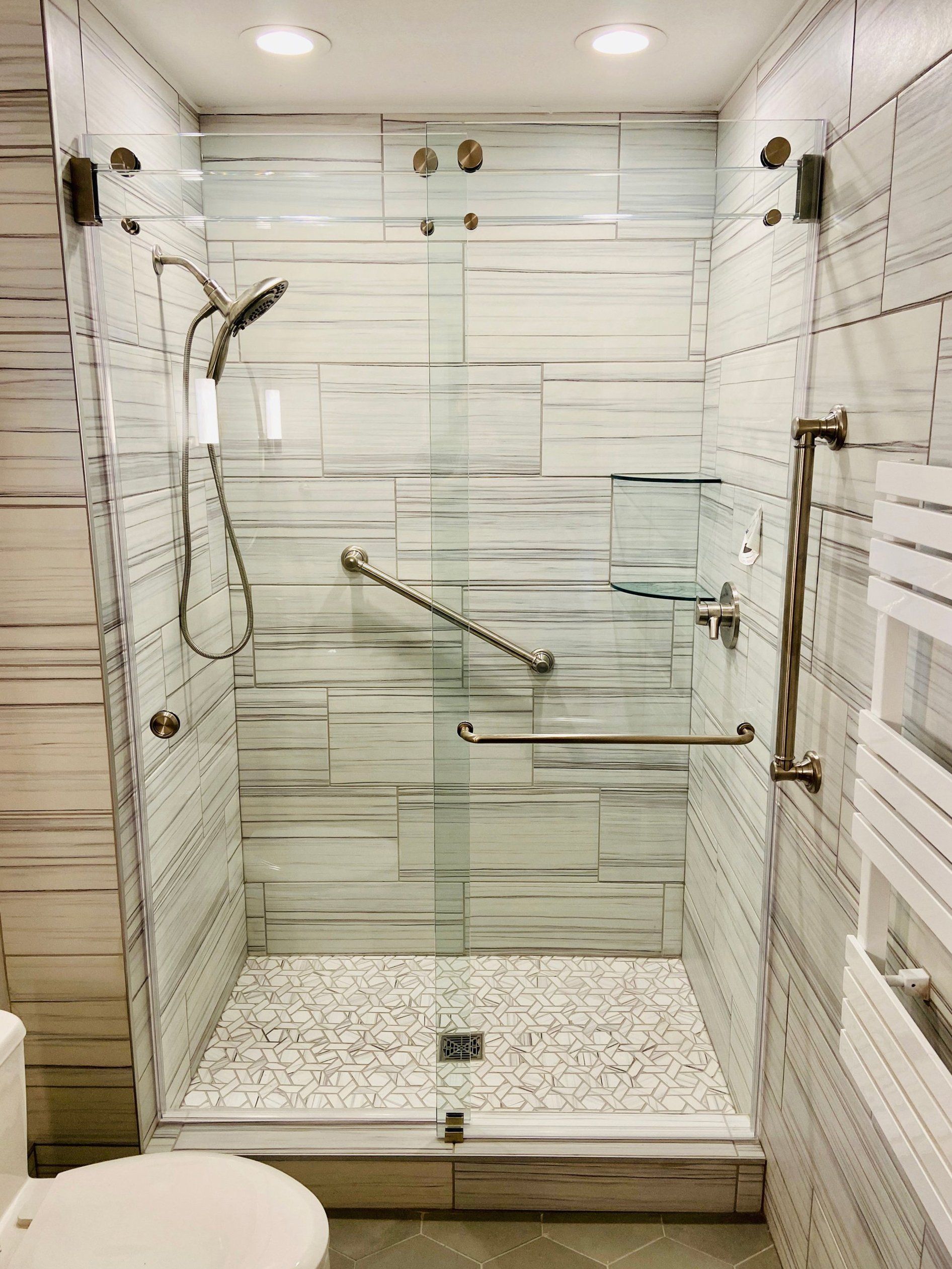 Walk-in shower with glass doors, white tile walls, grab bars, and built-in bench seat