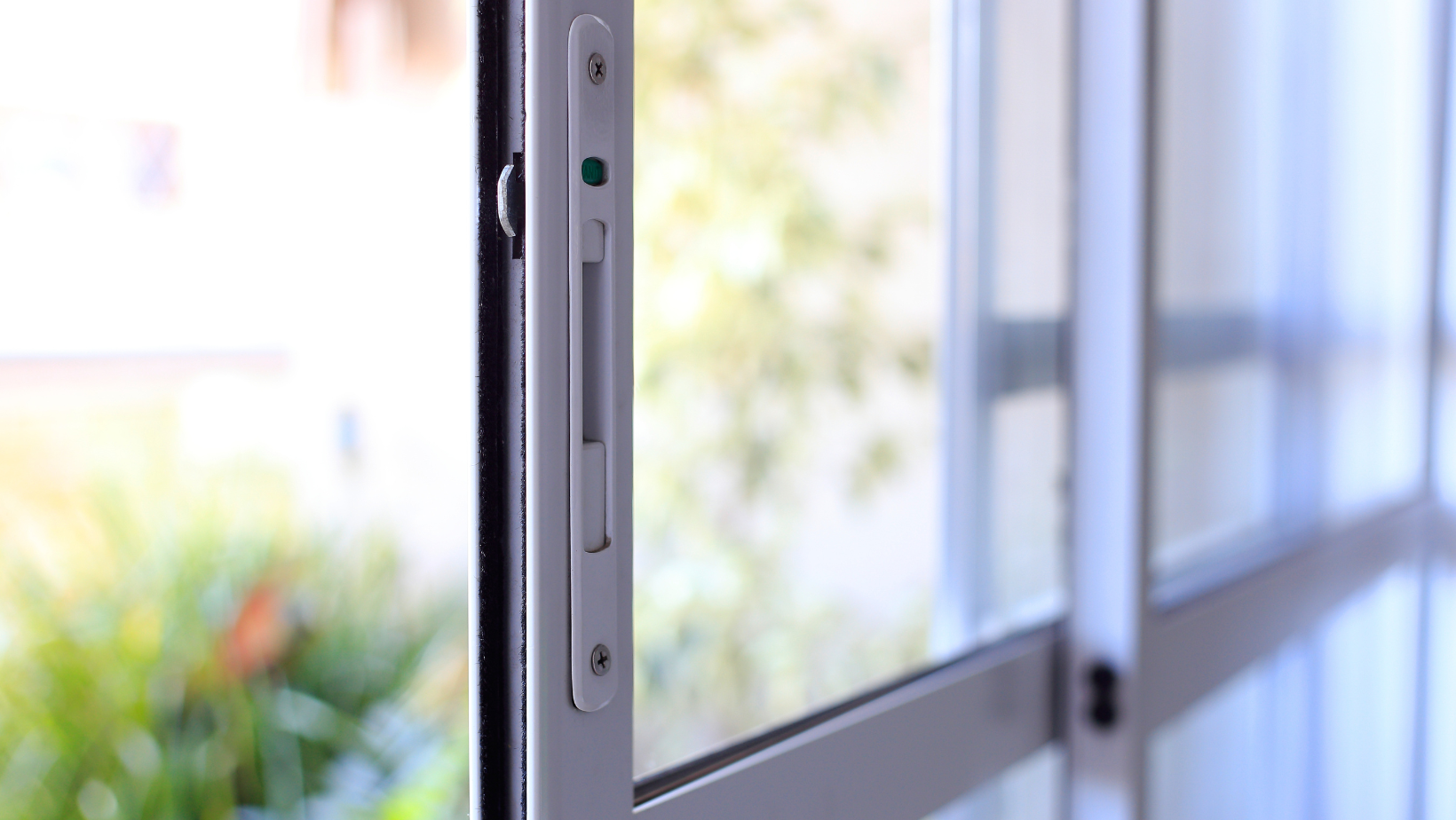 Close-up of a sliding glass door frame with a lock, next to a sunlit window and blurred greenery outside